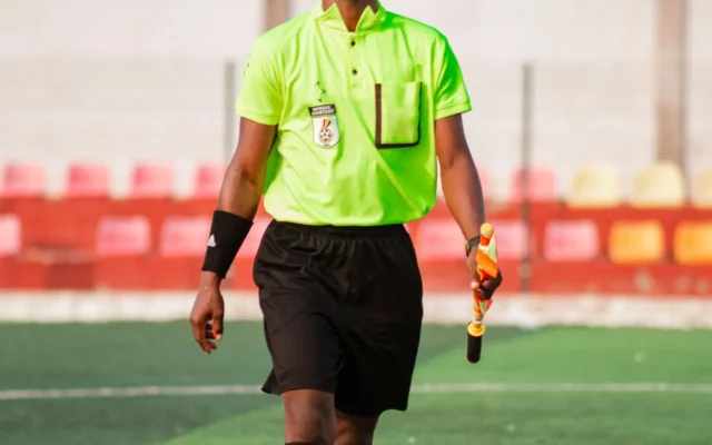 Arbitru asistent în echipament verde, cu fanion, pe un teren de fotbal.