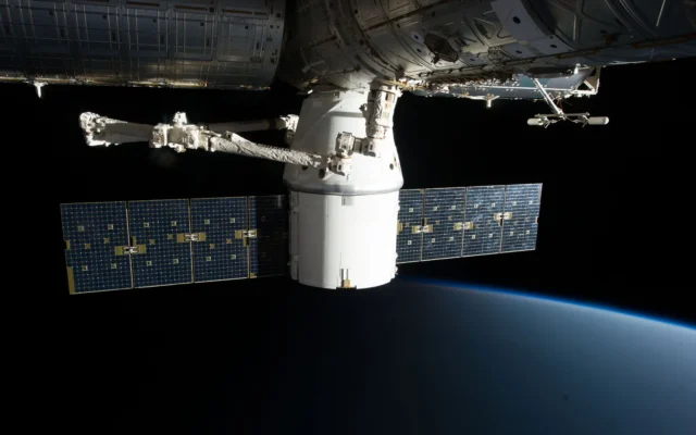 Capsulă spațială cu panouri solare și braț robotic lângă stația orbitală, deasupra Pământului