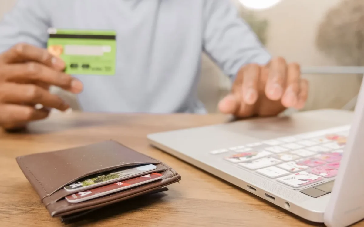 Mâini cu un card bancar verde, lângă un laptop și un portofel