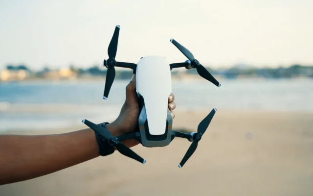 Mână care ține o dronă albă și neagră cu patru elice, lângă apă