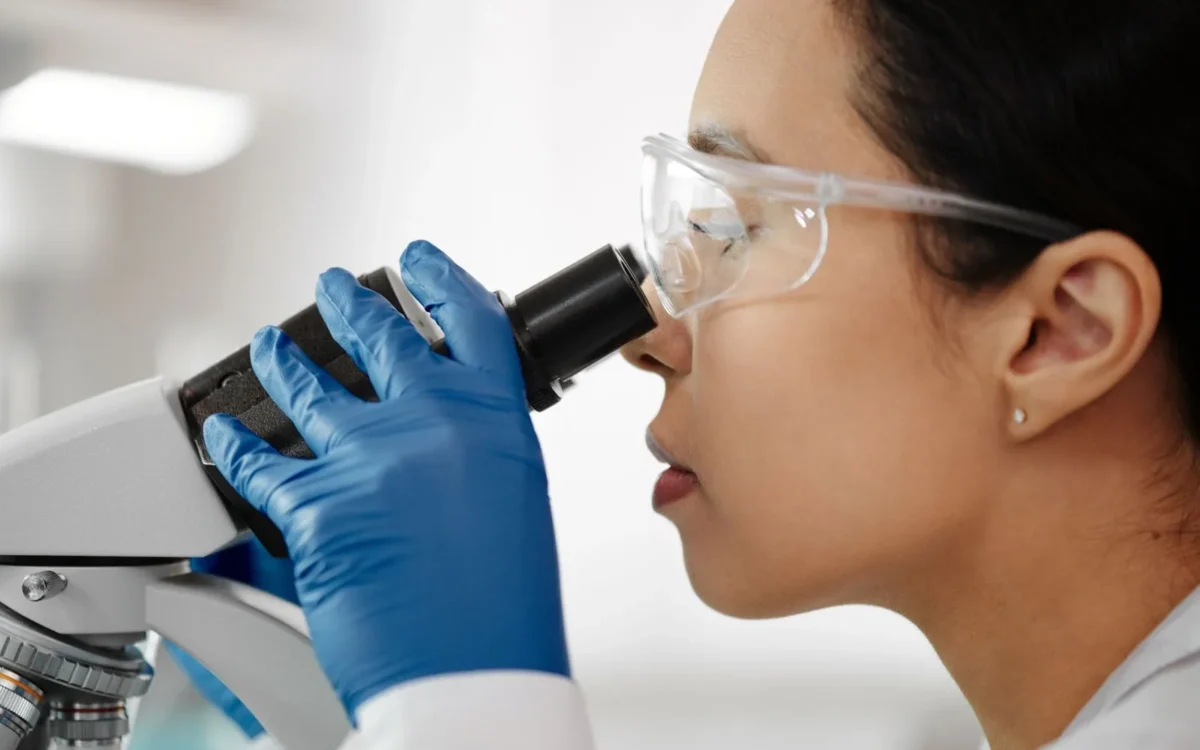 Femeie în halat alb și ochelari de protecție folosind un microscop de laborator