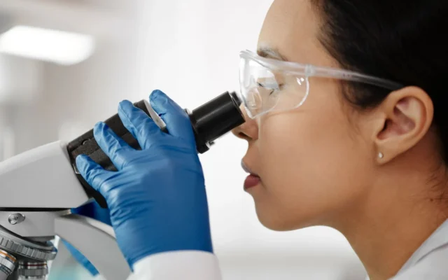 Femeie în halat alb și ochelari de protecție folosind un microscop de laborator