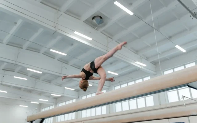 Gimnastă în echipament negru execută o mișcare pe bârnă, într-o sală de sport