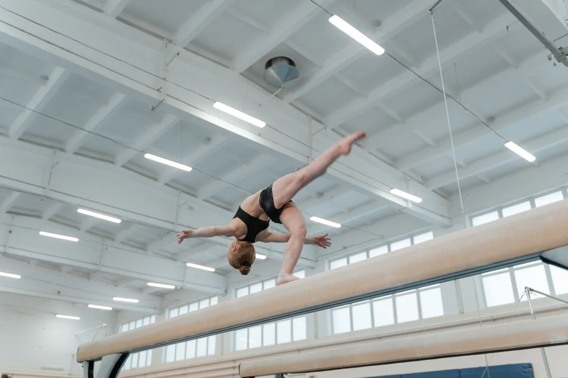 Gimnastă în echipament negru execută o mișcare pe bârnă, într-o sală de sport