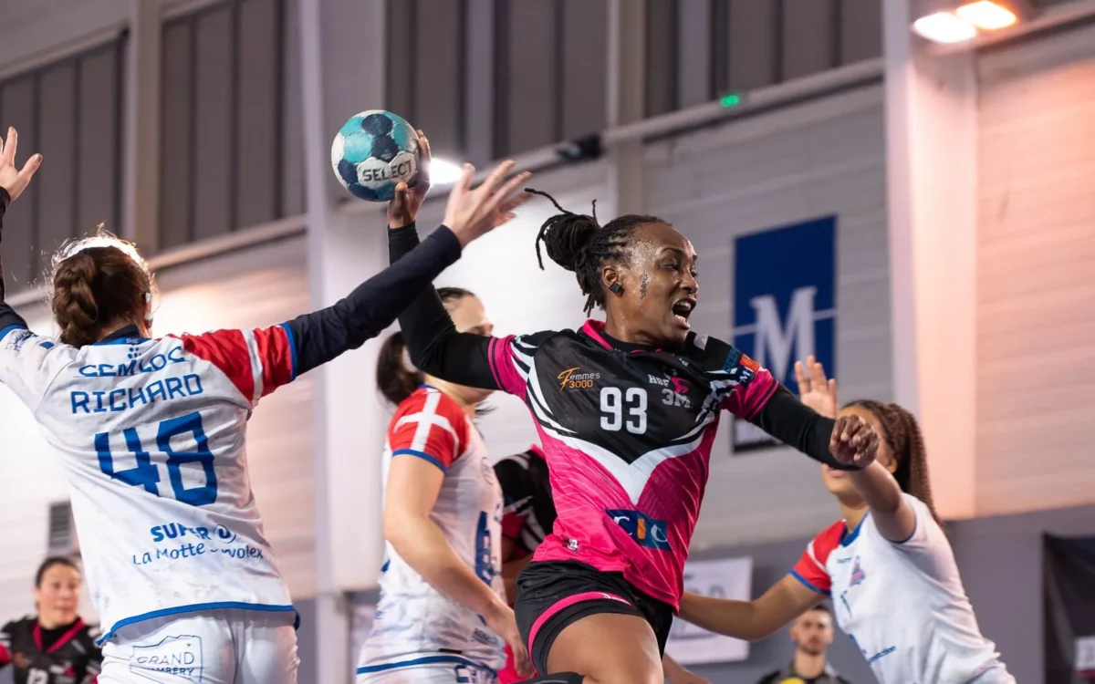Jucătoare de handbal în roz și negru, în săritură cu mingea, între adversare în alb