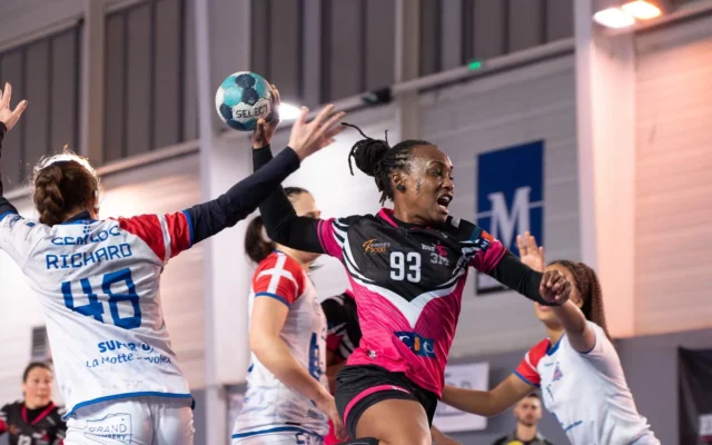 Jucătoare de handbal în roz și negru, în săritură cu mingea, între adversare în alb