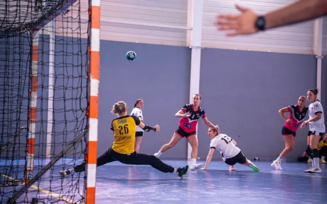 Jucătoare de handbal și un portar în fața porții, într-o sală de sport