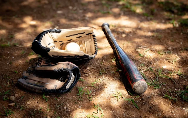 Două mănuși, o minge albă și o bâtă neagră de baseball, pe pământ