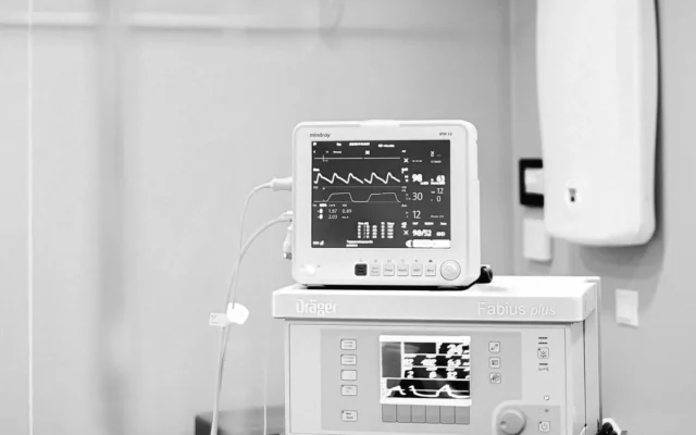 Monitor medical cu trasee și parametri vitali, lângă alte echipamente, în alb-negru