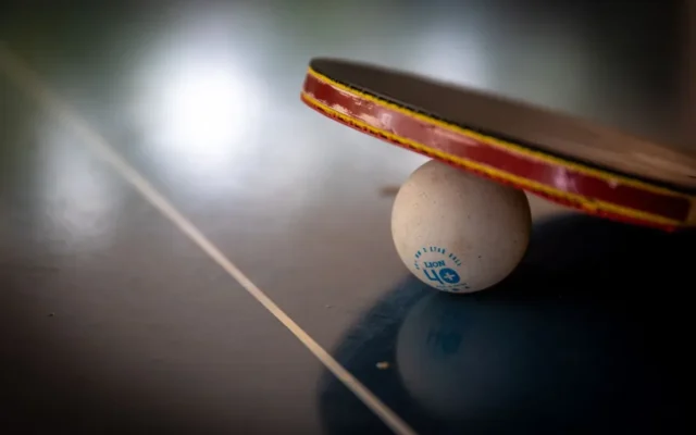 Paletă de tenis de masă sprijinită pe o minge albă, pe o masă întunecată