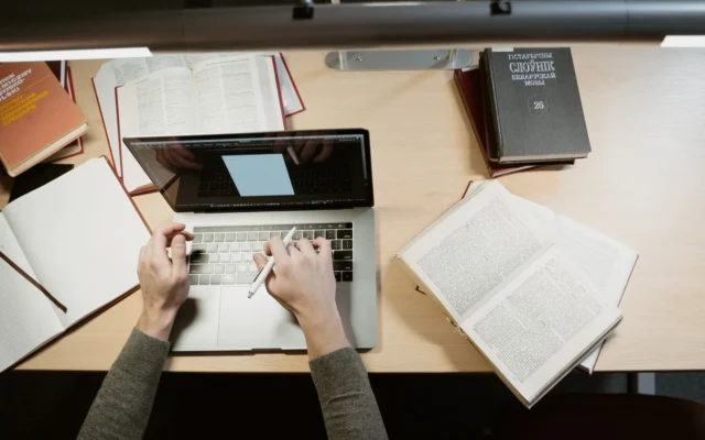 Persoană la laptop, lângă cărți și caiete