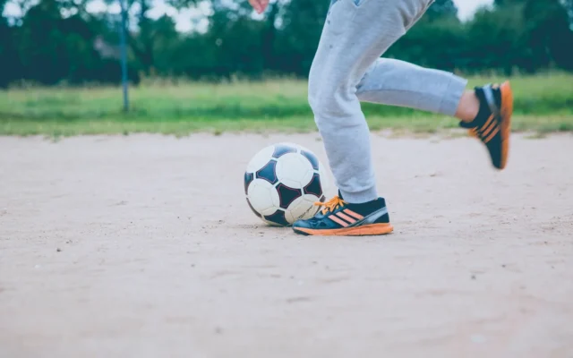 Persoană în pantaloni gri lovind o minge de fotbal pe un teren nisipos