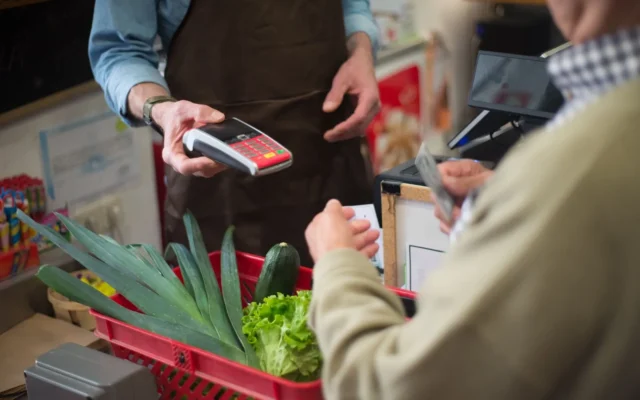 Client cu card bancar lângă terminalul de plată, cu un coș de legume la casa magazinului