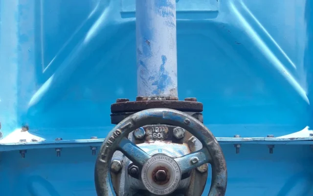 Robinet metalic pe o conductă albastră, în fața unui rezervor industrial