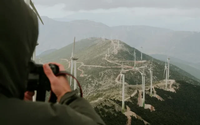 Persoană cu aparat foto în fața unor turbine eoliene amplasate pe coline