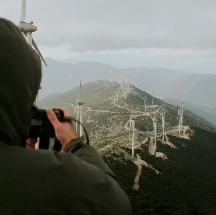 Persoană cu aparat foto în fața unor turbine eoliene amplasate pe coline